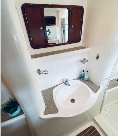 Compact boat bathroom (marine head) with white molded sink and countertop, chrome faucet, wood-framed mirrored cabinet, toiletries and small potted plant.