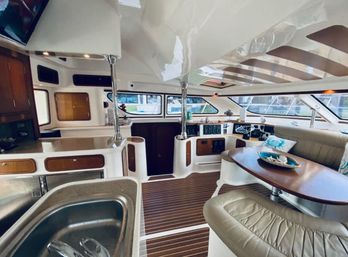 Sunlit modern yacht interior: helm station and wraparound windows with marina view, teak-style flooring, galley with stainless sink, curved leather seating and wooden dinette table with nautical decor.