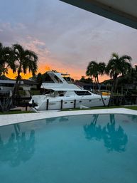 White motor yacht moored at a residential canal beside a turquoise backyard pool, silhouetted palm trees and a vivid orange sunset sky