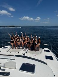 Group of women in black swimsuits and captain hats posing and cheering on the bow of a white yacht during a sunny boat party on open blue water