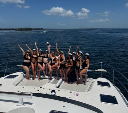 Group of women in black swimsuits and captain hats posing and cheering on the bow of a white yacht during a sunny boat party on open blue water