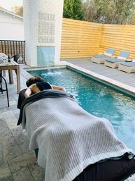 Person receiving an outdoor massage beside a narrow backyard plunge pool with a waterfall feature, stone patio, wooden privacy fence and lounge chairs on the deck
