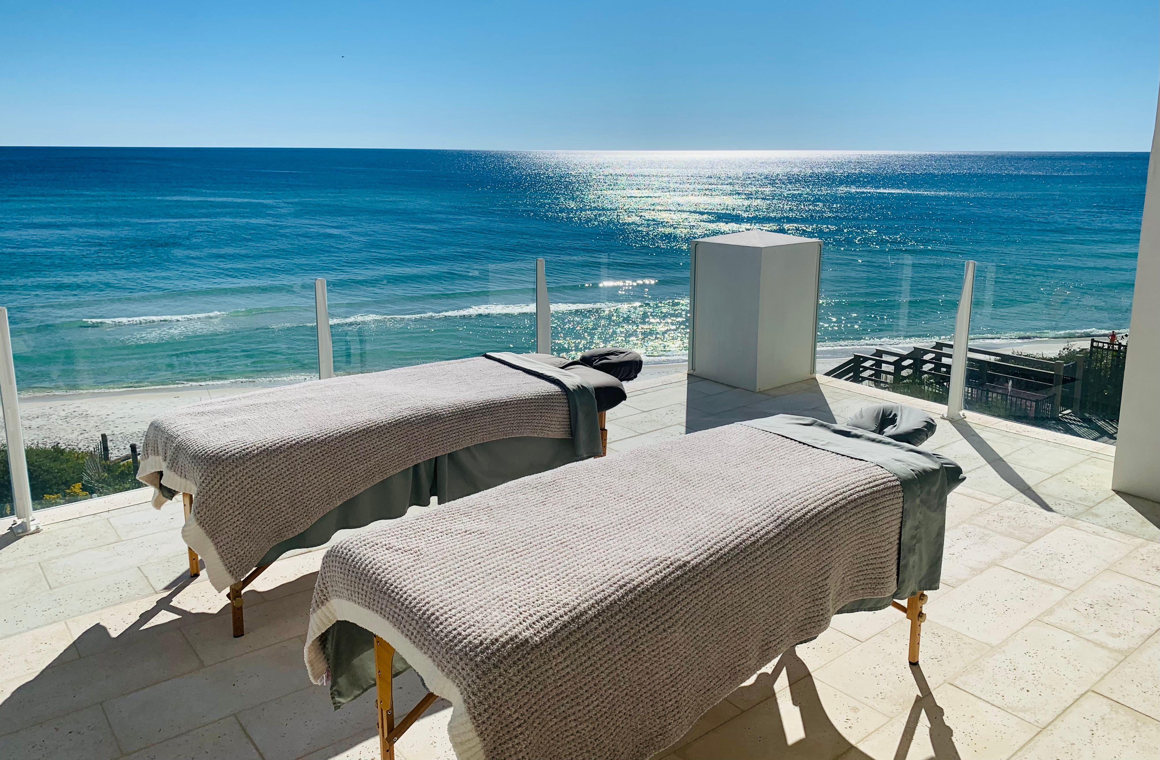 Two spa massage tables on a sunlit oceanfront terrace with glass railing overlooking sparkling turquoise water and a sandy beach.