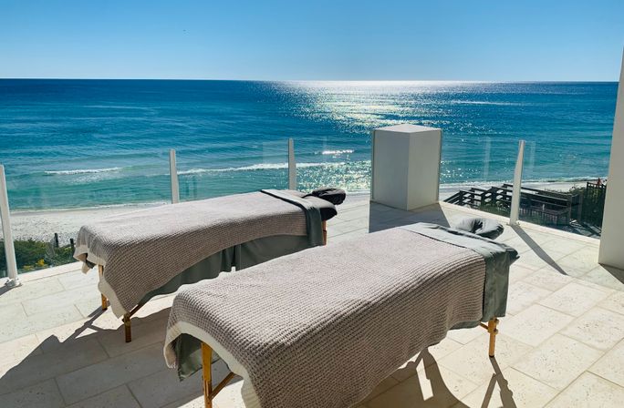 Two spa massage tables on a sunlit oceanfront terrace with glass railing overlooking sparkling turquoise water and a sandy beach.