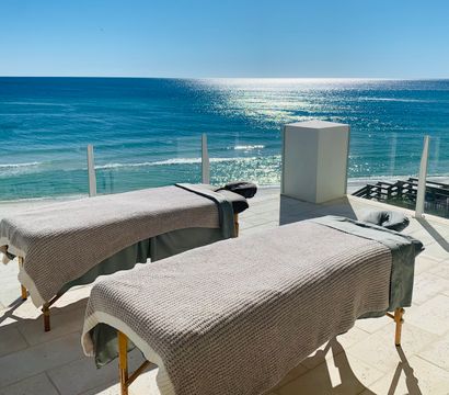 Two spa massage tables on a sunlit oceanfront terrace with glass railing overlooking sparkling turquoise water and a sandy beach.