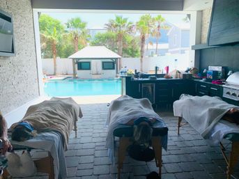 Poolside outdoor spa scene with three massage tables and towel-covered clients in an open-air kitchen area overlooking a turquoise pool, palm trees and a cabana in a sunny tropical backyard.