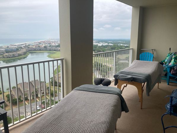 Two massage tables with gray blankets on a high-rise balcony overlooking a lagoon, rows of beachfront homes and the distant ocean under an overcast sky