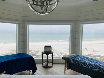 Beachfront spa treatment room with two massage tables and a towel warmer on a small round table, panoramic windows framing white sand dunes and calm turquoise ocean under a cloudy sky.