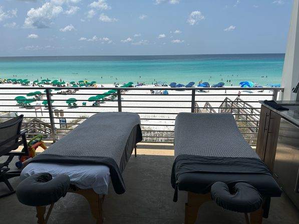 Beachfront spa balcony with two massage tables facing turquoise ocean and white-sand beach dotted with green and blue umbrellas under a sunny sky