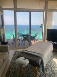 Massage table in a bright beachfront condo living room, sliding glass doors opening to a balcony with table and chairs and sweeping turquoise ocean view.