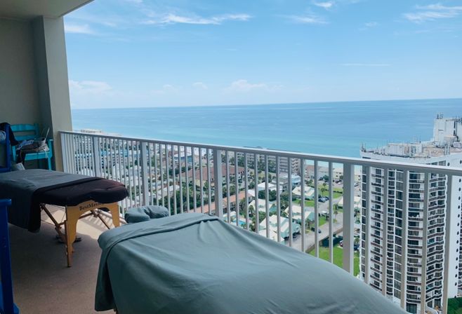 High-rise balcony spa setup with two portable massage tables and teal linens overlooking turquoise ocean, beachfront condos and a sunny coastal skyline