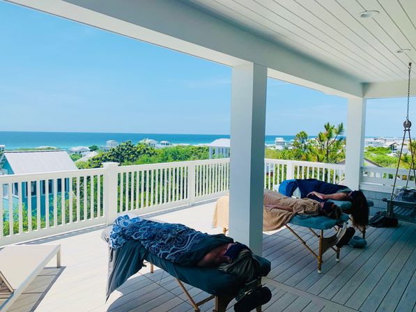 Covered beachfront balcony with white railing and ocean view, two massage tables with people relaxing and coastal homes under a bright blue sky