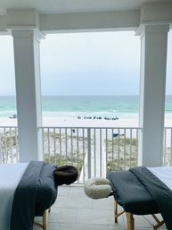 Relaxing setup with two massage tables on a covered beachfront balcony overlooking white sand, turquoise ocean, a wooden boardwalk to the shore, and a distant row of blue beach chairs.