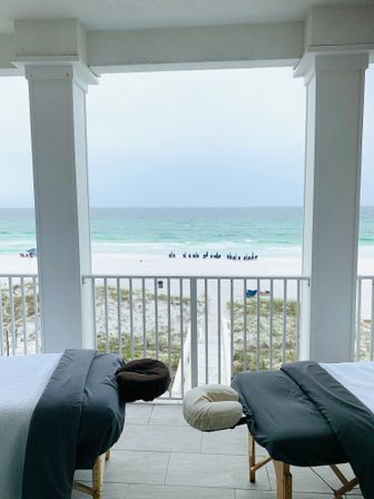 Relaxing setup with two massage tables on a covered beachfront balcony overlooking white sand, turquoise ocean, a wooden boardwalk to the shore, and a distant row of blue beach chairs.