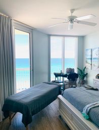 Sunlit oceanfront bedroom in a beachfront condo with a massage table and bed facing large windows and a balcony overlooking turquoise ocean and sandy beach.