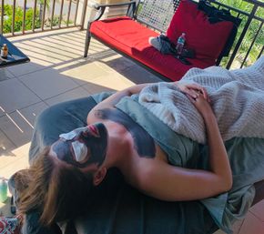 Person relaxing on a sunny balcony receiving a charcoal facial with under-eye patches, wrapped in a cozy blanket on an outdoor lounge.