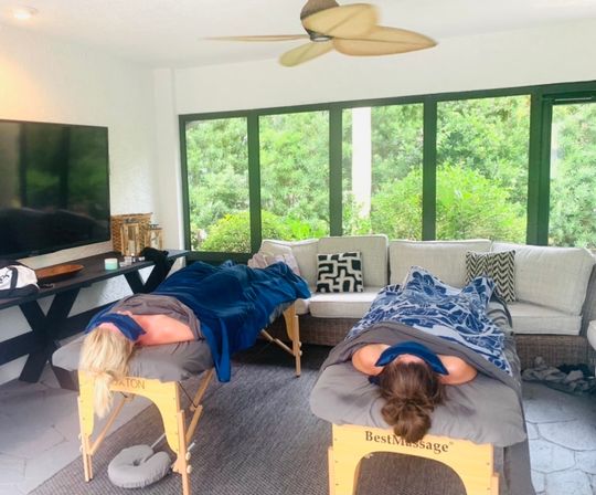 Relaxing sunroom spa setup with two massage tables and clients under blankets, large windows overlooking a lush garden, ceiling fan and wicker sofa