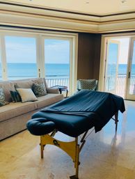 Beachfront living room with a portable massage table draped in dark cloth, beige sofa and patterned chair beside large windows and balcony doors framing a blue ocean horizon.