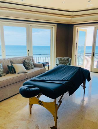 Beachfront living room with a portable massage table draped in dark cloth, beige sofa and patterned chair beside large windows and balcony doors framing a blue ocean horizon.