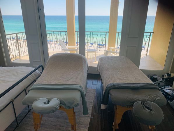 Two massage tables with gray covers side-by-side in a beachfront room, facing a sunlit balcony and turquoise ocean with sandy beach and blue umbrellas — spa-ready, relaxing ocean-view setup.