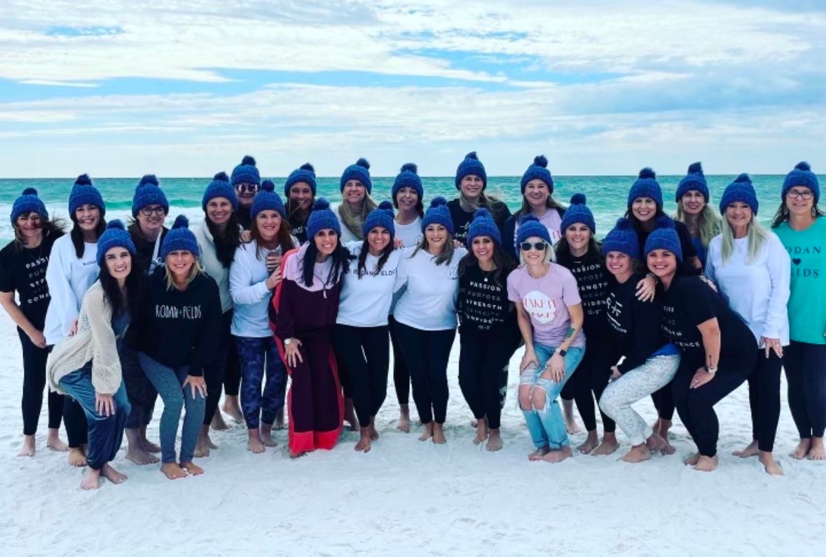 Smiling group of women wearing matching blue knit beanies, barefoot on a white-sand beach with turquoise ocean and cloudy sky.