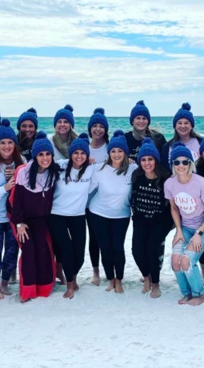 Smiling group of women wearing matching blue knit beanies, barefoot on a white-sand beach with turquoise ocean and cloudy sky.