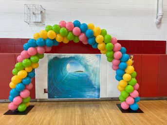 Colorful balloon arch of pink, blue, yellow and green balloons framing an ocean wave backdrop inside a school gymnasium with red padded walls and hardwood floor