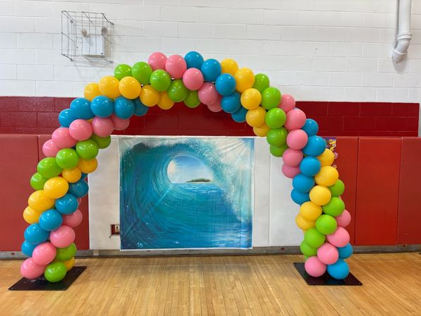Colorful balloon arch of pink, blue, yellow and green balloons framing an ocean wave backdrop inside a school gymnasium with red padded walls and hardwood floor