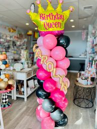 Vibrant balloon column of pink, black and gray balloons topped by a yellow Birthday Queen crown foil balloon with gold letter balloons spelling a name, displayed as a party shop decoration.