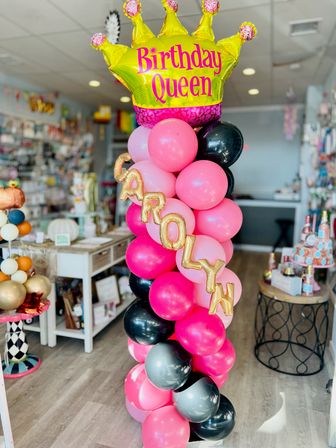 Vibrant balloon column of pink, black and gray balloons topped by a yellow Birthday Queen crown foil balloon with gold letter balloons spelling a name, displayed as a party shop decoration.
