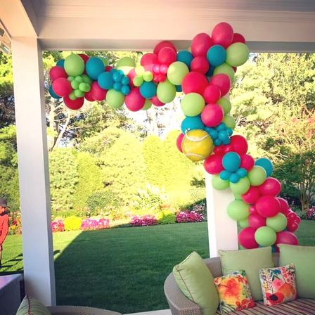 Bright pink, teal and lime green balloon garland draped over a white backyard patio column above a wicker sofa with floral cushions, overlooking a manicured lawn and garden beds