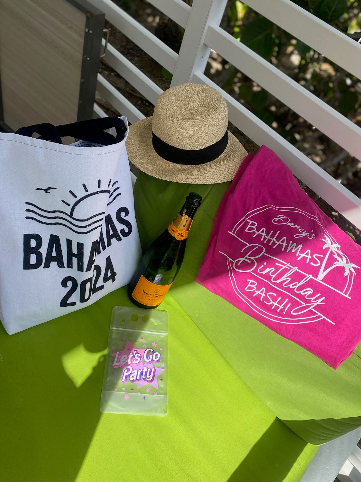 Bahamas vacation flatlay on a lime-green cushion: straw sun hat, white tote bag reading Bahamas 2024, pink Bahamas Birthday Bash t-shirt, a bottle of sparkling wine, and a 'Let's Go Party' pouch.