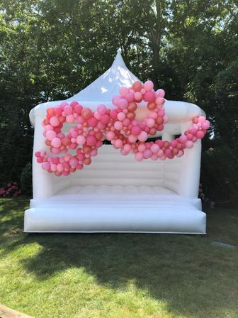 White inflatable bounce house tent on a sunlit backyard lawn, draped with cascading pink balloon garlands in various shades for party decor