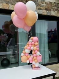 Giant number 4 display filled with pastel pink, peach, and white balloons, topped with a matching pastel balloon bouquet on a patio table in front of a stone facade and sliding glass doors — backyard birthday party decoration.