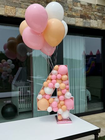 Giant number 4 display filled with pastel pink, peach, and white balloons, topped with a matching pastel balloon bouquet on a patio table in front of a stone facade and sliding glass doors — backyard birthday party decoration.