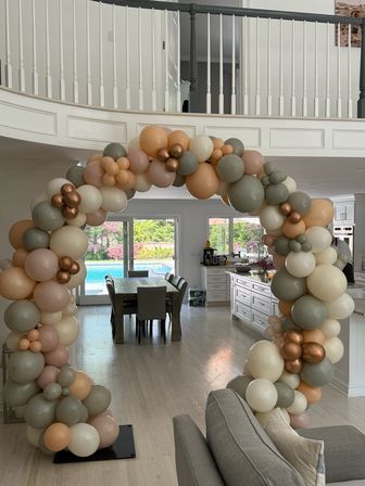 Neutral pastel balloon arch framing an open-plan modern living space with dining table and sliding doors to a backyard pool — cream, peach, sage and metallic copper balloons.