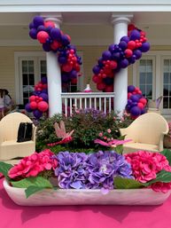 Bright pink and purple balloon garlands spiraling around white porch columns with a matching pink and purple hydrangea centerpiece, wicker chairs and floral patio décor for an outdoor party