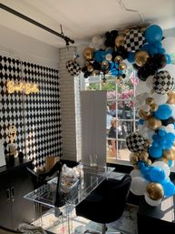 Bright studio/event space with black-and-white diamond wallpaper, clear acrylic desk and black swivel chair under a modern glass-bulb chandelier, large blue, gold, black, white and checkered balloon arch framing a street-facing window, and a small neon cursive sign on the wall — party decor and shopfront setup.