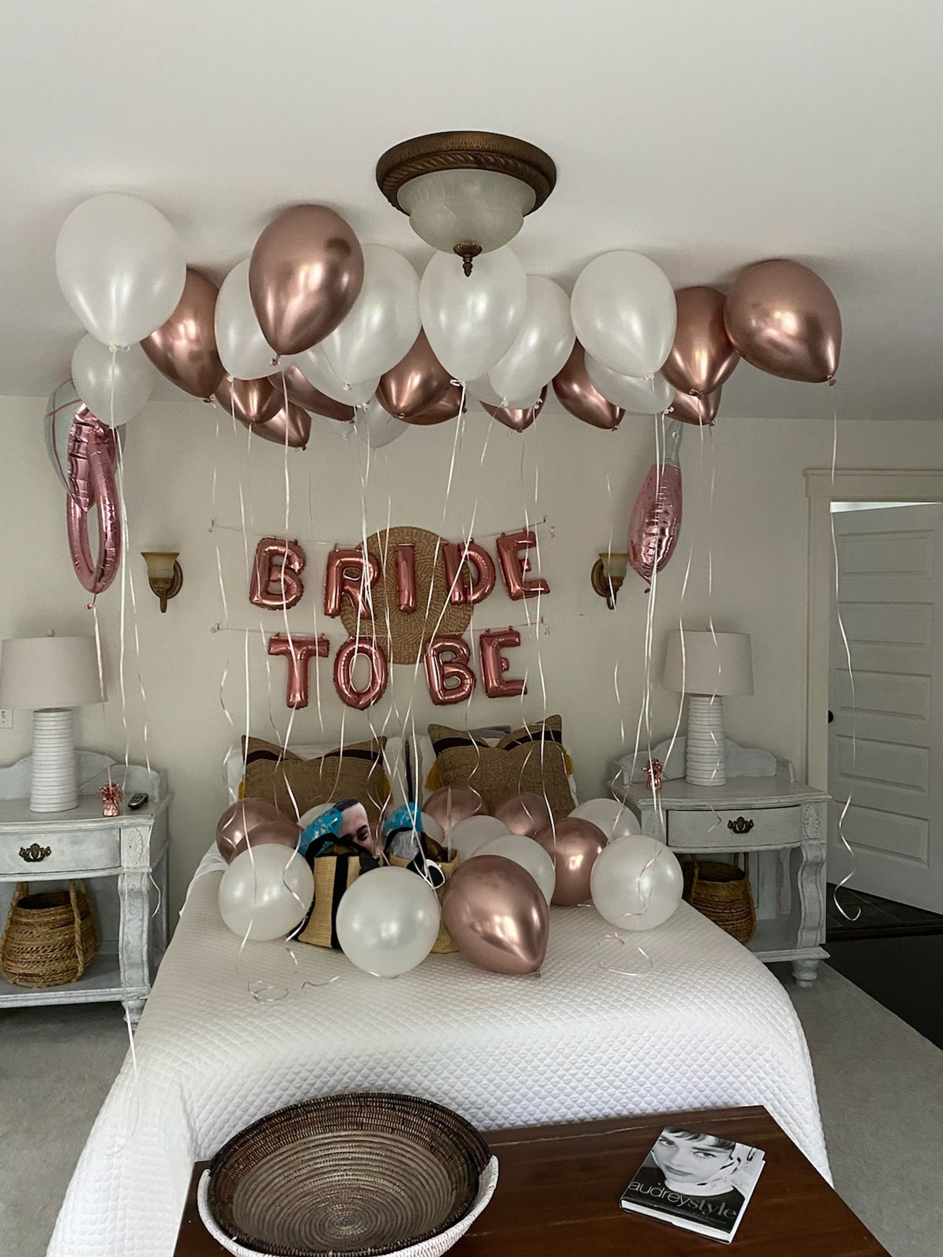 Cozy neutral bedroom staged for a bridal shower with rose gold and white helium balloons floating above a bed and a rose-gold "BRIDE TO BE" balloon banner, bedside tables with lamps and woven baskets