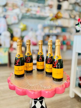 Five mini champagne bottles with yellow labels arranged on a pink scalloped pedestal in a colorful boutique gift shop display