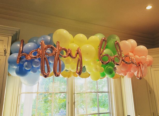 Pastel rainbow balloon garland with rose-gold script 'happy bday' balloons hung above a bright window in a home