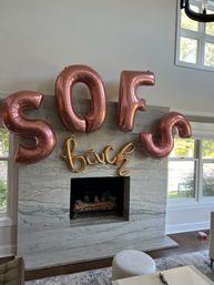 Rose-gold foil letter balloons reading "SOFS" with a smaller gold script balloon saying "back" hung above a marble fireplace in a bright living room.