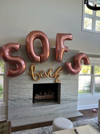 Rose-gold foil letter balloons reading "SOFS" with a smaller gold script balloon saying "back" hung above a marble fireplace in a bright living room.