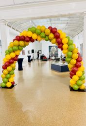 Festive red, yellow, and green balloon arch framing a bright indoor art gallery with arched skylight, exhibition walls and a small stage setup in the background.