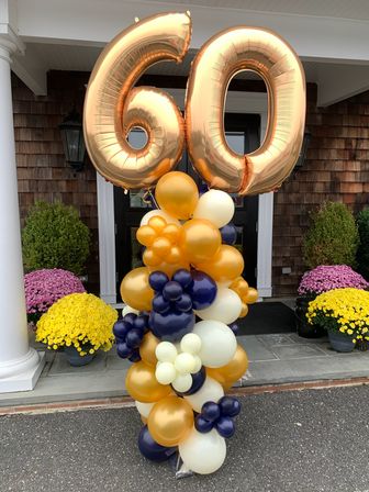 Front porch 60th birthday balloon display with large gold foil '60' numbers atop a navy, gold and ivory balloon column, flanked by yellow and pink potted mums at a house entrance.