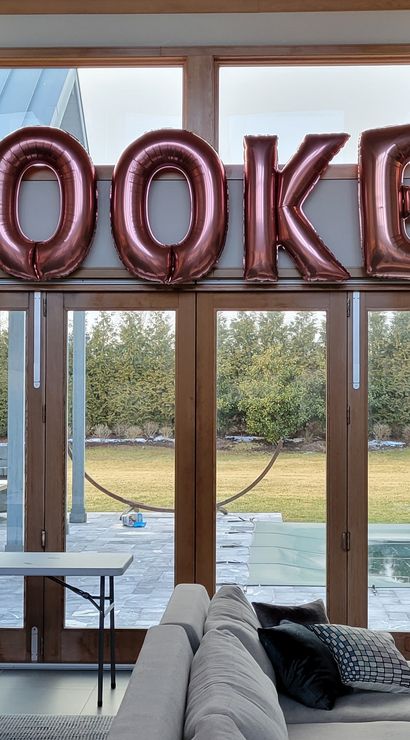 Contemporary living room with floor-to-ceiling patio doors overlooking a backyard pool, decorated for a party with rose-gold metallic letter balloons spelling a name, gold cursive balloons, and a peach-white-gold balloon garland draped over the doorway.
