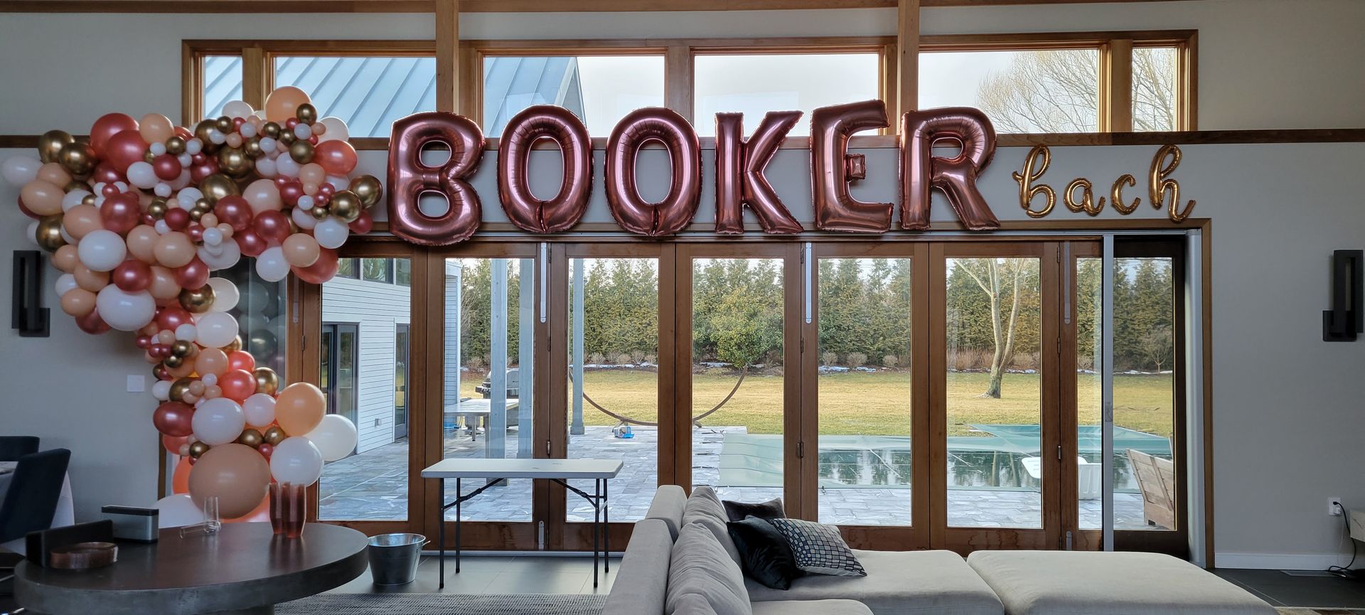 Contemporary living room with floor-to-ceiling patio doors overlooking a backyard pool, decorated for a party with rose-gold metallic letter balloons spelling a name, gold cursive balloons, and a peach-white-gold balloon garland draped over the doorway.