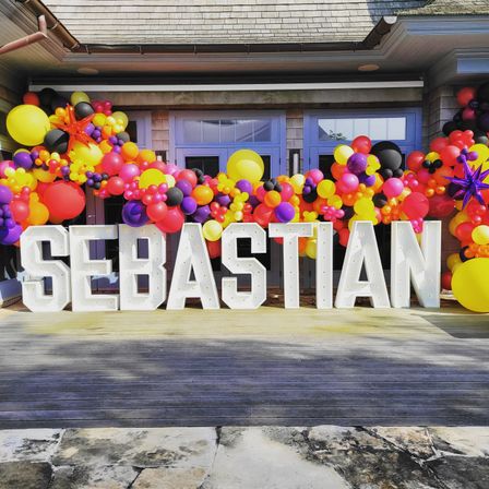 Bright outdoor party scene with oversized white illuminated marquee letters forming a name on a wooden deck front porch, framed by a vivid balloon garland in yellow, pink, purple, orange and black.
