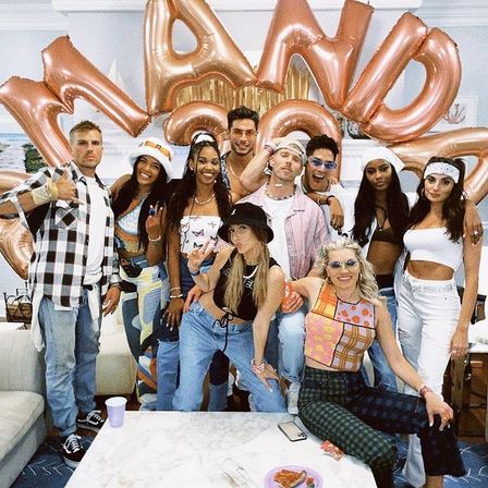 Smiling group of friends posing in a living-room party with rose-gold letter balloons, retro-inspired outfits, and snacks on a coffee table.