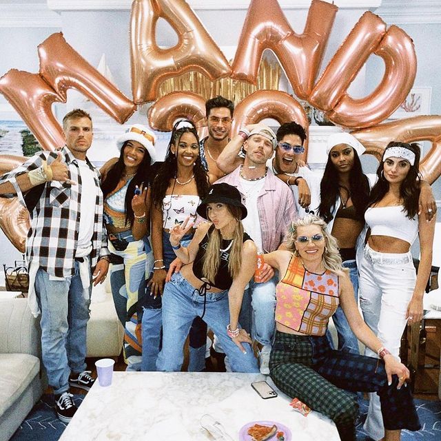 Group of friends posing at an indoor house party with rose-gold letter balloons, Y2K-inspired retro outfits, casual drinks and playful poses — lively living-room celebration photo.